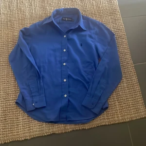 Blå skjorta från Ralph Lauren - Klassisk blå skjorta från Ralph Lauren med lång ärm och knappar framtill. Skjortan har en stilren design med liten broderad logga på bröstet och är tillverkad i ett mjukt bomullsmaterial. Perfekt för en clean och tidlös look.