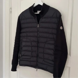 Svart cardigan från Moncler - Säljer en svart pufferjacka från Moncler med quiltad front och ärmar i stickad kvalitet. Jackan har dragkedja framtill och Moncler-logga på ena ärmen. Perfekt för kyliga dagar och snygg till vardags.