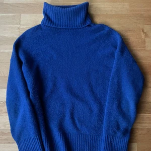 Blå polotröja från & Other Stories - Säljer en blå polotröja från & Other Stories Paris Atelier. Tröjan är stickad med hög polokrage och breda ribbade muddar vid ärmslut och nederkant. Perfekt för kyliga dagar och enkel att styla till olika outfits.