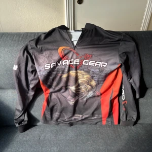 Svart och röd långärmad tröja från Savage Gear - Sportig långärmad tröja från Savage Gear med dragkedja vid halsen. Svart bas med röda och orange detaljer samt stort fiskmotiv framtill. Tryck med logga och texten 'Water Wolf' på ärmen. Perfekt för fiske eller utomhusaktiviteter. Väldigt luftigt material! :)