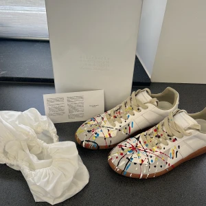 Maison Margiela sneakers med färgstänk - Unika sneakers från Maison Margiela med vit bas och färgstänk i rött, blått, gult och svart. Skorna har snörning, rund tå och en brun gummisula. Tillverkade i skinn. Nypris är ca 9300 kr, vårt pris är 3199! Skriv bara om ni har frågor!