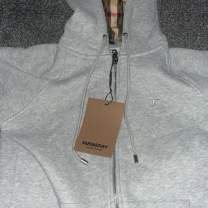 Ljusgrå hoodie från Burberry - Säljer en stilren ljusgrå hoodie från Burberry med dragkedja och klassiskt rutigt foder i huvan. Tröjan har långa ärmar, justerbar huva med snören och subtilt broderad logga på bröstet. Perfekt för en avslappnad och trendig look. Pris Kan diskuteras