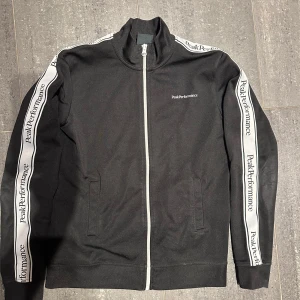 Svart träningsjacka från Peak Performance - Svart träningsjacka med vit dragkedja och vita ränder längs ärmarna där det står Peak Performance. Jackan har hög krage och två fickor. 