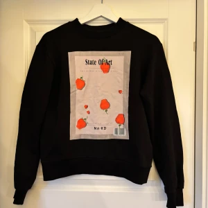 Svart sweatshirt med jordgubbstryck från NA-KD - Cool svart sweatshirt från NA-KD med stort tryck på framsidan med texten 'State Of Art' och flera röda jordgubbar. Rund halsringning och ribbade muddar vid ärmslut och nederkant. Perfekt för en avslappnad stil.