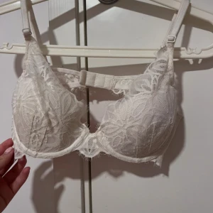 Vit spets-bh med blommönster - Säljer en vit bh med vackert blommigt spetsmönster och justerbara axelband. Bh:n har bygel och dekorativ spetsdetalj framtill. Perfekt för dig som gillar romantiska detaljer. Storlek 75B, använt fåtal gånger, i mycket bra skick!