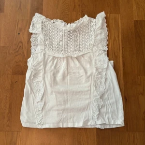 Vit spetsdetaljerad linnetopp från MNG - Superfin vit linnetopp från MNG med broderade spetsdetaljer och volanger längs axlarna. Toppen är ärmlös och har vackert mönstrad spets framtill samt en liten knapp i nacken. Perfekt för en somrig look!