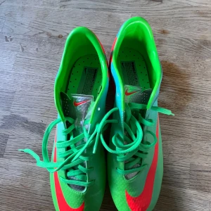 Nike Mercurial fotbollsskor i neonfärg - Säljer ett par Nike Mercurial fotbollsskor i grön neon med röda detaljer och snörning. Skorna har platt sula med dobbar för bra grepp på planen och är tillverkade i syntetmaterial. Perfekta för dig som vill sticka ut på fotbollsplanen! Helt oanvända! kom med bud