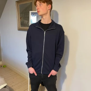 Mörkblå tröja med dragkedja - Säljer en stilren mörkblå tröja med dragkedja. Perfekt för en avslappnad look. Tröjan har långa ärmar och en hög krage som kan bäras uppvikt eller nedvikt. Passar bra till jeans eller chinos. Kommer från 157