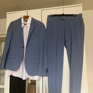 Ljusblå kostym från Dressmann - Säljer en ljusblå kostym från Dressmann i slim fit. Kavajen har två knappar framtill och klassiska fickor, stl S46. Byxorna är i matchande färg och har storlek 31-30. Perfekt för dig som vill ha en stilren och modern look.