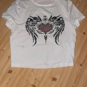 Vit croppad t-shirt med tribaltryck - Säljer en vit croppad t-shirt med ett stort svart tribaltryck och ett rött hjärta på bröstet. T-shirten har korta ärmar och en klassisk rund halsringning. Perfekt för dig som gillar edgy och unik stil.