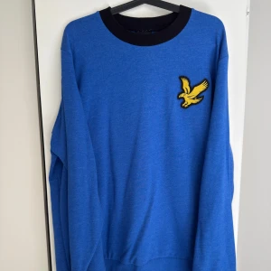 Blå tröja från Lyle & Scott med gul fågel XL - Säljer en blå långärmad tröja från Lyle & Scott med svart ribbad krage och deras klassiska gula fågel-logga på bröstet. Enkel och stilren design som passar perfekt till jeans eller chinos.