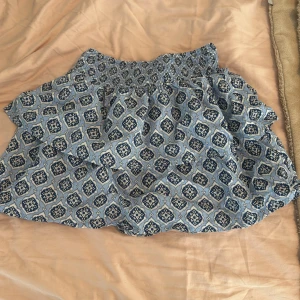 Blåmönstrad kortkjol från Gina Tricot - Supersöt kortkjol från Gina Tricot med blått och vitt mönster. Kjolen har smock i midjan och volangdetalj som ger en luftig och skön känsla. Perfekt för varma dagar och enkel att matcha med en enkel topp.