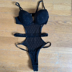 Svart mesh body med strassdetaljer - Säljer en svart body med vadderade kupor och meshmaterial. Den har glittriga strassdetaljer, smala axelband och utskurna partier i midjan. Perfekt för att skapa en statement-look.