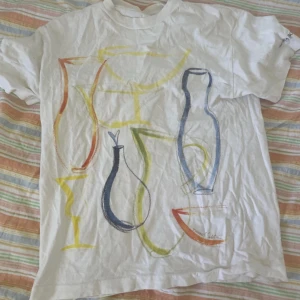 Vit t-shirt med färgglatt konstmotiv från Kosabda - Vit t-shirt med färgglatt abstrakt tryck av vaser och glas i blått, gult, rött och grönt. Klassisk passform och rund hals. Perfekt för dig som gillar unika och konstnärliga plagg.