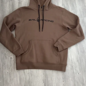 Brun hoodie från Sail Racing - Säljer en brun hoodie från Sail Racing i storlek L. Tröjan har svart tryck på bröstet, huva med svarta snören och en stor magficka. Perfekt för dig som gillar en avslappnad och sportig stil.Nypris är 1300kr men säljer den för 900kr! En liten liten fläck på högra axeln som går säkert att tvätta bort 