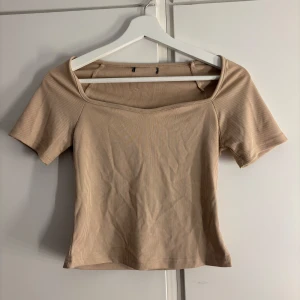 Beige ribbad topp med fyrkantig ringning - Säljer en beige ribbad topp med korta ärmar och snygg fyrkantig ringning. Toppen har en tight passform och är tillverkad i ett mjukt, stretchigt material. 