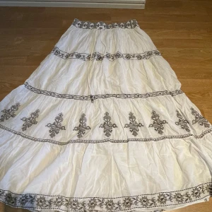 Vit långkjol med broderi från Zara - Säljer en vit långkjol från Zara med vackra svarta broderade blommor och mönster längs kjolen. Perfekt för sommarens alla tillfällen!