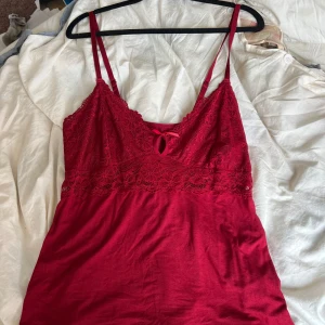 Röd spetslinnetopp med rosett - Säljer en röd linnetopp med smala axelband och vacker spets upptill. Toppen har en liten rosett framtill och blommigt spetsmönster över bysten. Perfekt för dig som gillar feminina detaljer och en romantisk look.
