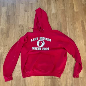 Röd hoodie från Hanes med tryck - Röd hoodie från Hanes med tryck på bröstet: 'Lady Indians Water Polo' och en vattenpolo-boll, samt 'Fullerton' tryckt på ryggen. Klassisk känguruficka och huva med dragsko. Perfekt för dig som gillar sportig stil. Lite målarfärg stänk på ena ärmen