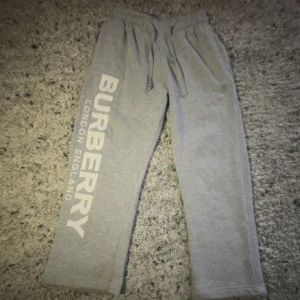 Grå mjukisbyxor från Burberry - Säljer ett par grå mjukisbyxor från Burberry med stort vitt tryck längs ena benet och broderad logga vid fickan. Byxorna har dragsko i midjan och är tillverkade i mjukt material för maximal komfort.