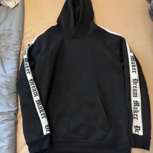 Svart hoodie med text på ärmar från Manfinity - Svart hoodie från Manfinity med vita ränder längs ärmarna där det står 'Dream Maker' i svart text. Klassisk känguruficka framtill och huva. Perfekt för en avslappnad stil.