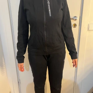 Svart zip-jacka från EA7 Emporio Armani - Säljer en svart jacka från EA7 Emporio Armani med dragkedja framtill och hög krage. Jackan har en sportig look och är tillverkad i ett mjukt syntetmaterial. Perfekt för en avslappnad stil.
