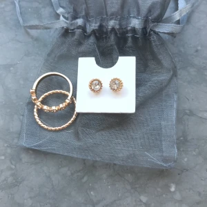Set med guldfärgade ringar och örhängen - Säljer ett set med två guldfärgade ringar och ett par matchande örhängen. Ena ringen har glittrande stenar runtom och den andra har en enkel, räfflad design. Örhängena är små studs med gnistrande stenar. Perfekt för dig som gillar stilrena accessoarer.