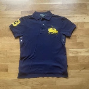 Ralph lauren piké - Säljer en klassisk mörkblå pikétröja från Polo Ralph Lauren med gul broderad logga på bröstet och siffra på ärmen. Tröjan har korta ärmar, krage och knappar framtill. Perfekt för en sportig och stilren look.