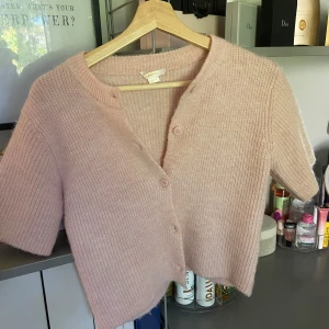 Ljusrosa stickad kofta med kort ärm - Ljusrosa stickad kofta med korta ärmar och v-ringning. Koftan har ribbad struktur och knäpps med tre knappar framtill❤️