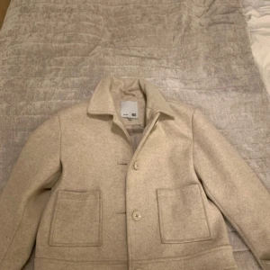 Barn Beige ullkappa från 157 - Snygg beige kappa från 157 med klassisk krage och stora framfickor. Jackan har knäppning framtill med knappar och är tillverkad i ett mjukt ullmaterial. Perfekt för dig som vill ha en stilren och tidlös look.