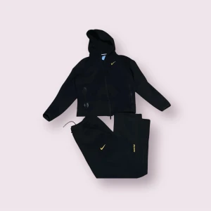 Svarta mjukisbyxor från Nike x Nocta - Svarta mjukisbyxor från Nike x Nocta med gul logga på benet och dragkedja vid fickan. Byxorna har en avslappnad passform och är perfekta för en sportig och chill stil.