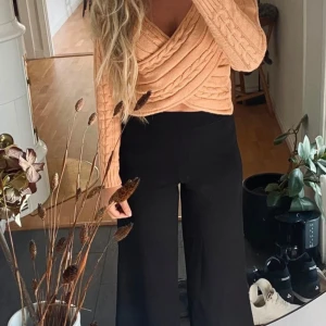 Stickad omlottopp i aprikos - Superfin stickad omlottopp i aprikos med kabelstickat mönster. Tröjan har långa ärmar och croppad passform, perfekt att styla med höga byxor. Snygg och trendig detalj med omlott över bysten.