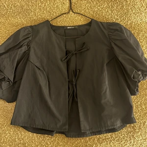 Svart blus med puffärmar och knytning - Säljer en svart blus med stora puffärmar och två dekorativa knytningar framtill. Blusen har en lös passform och är perfekt för dig som gillar unika detaljer och volym i ärmarna.