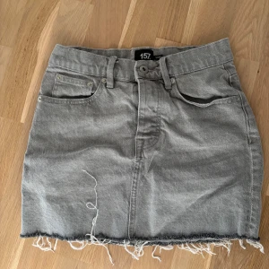 Grå jeanskjol med rå kant - Grå jeanskjol med klassiska fickor fram och bälteshällor. Kjolen har en rå, fransig kant nedtill för en avslappnad look. Perfekt att styla med en enkel t-shirt eller hoodie.