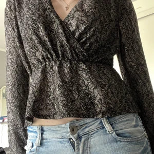 Svart omlottopp med mönster - Säljer en svart topp med silvrigt mönster och långa ärmar. Toppen har v-ringning och markerad midja som ger en snygg siluett. Perfekt att styla med jeans eller kjol.