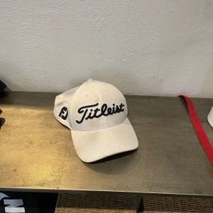 Vit keps från Titleist - Snygg vit keps från Titleist med svart broderad logga framtill och FJ-märke på sidan. Klassisk böjd skärm. Perfekt för golf eller vardagsstil. Den ser smutsig ut på bilden men kommer helt ny tvättad, ren och fräsch. 