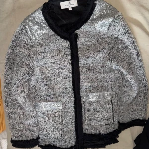 Glittrig silver kavajjacka med svarta detaljer - Säljer en glittrig silverfärgad kavajjacka från Rave Review med svarta kanter och två fickor framtill. Jackan har en rak passform och är långärmad. Perfekt för dig som vill sticka ut med en unik och trendig look.