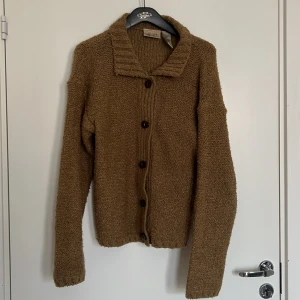 Brun stickad kofta från Kathie Lee - Mysig brun stickad kofta från Kathie Lee med stora knappar framtill och krage. Koftan har lång ärmad och är tillverkad i 100% akryl. Perfekt för lager-på-lager under kyligare dagar.