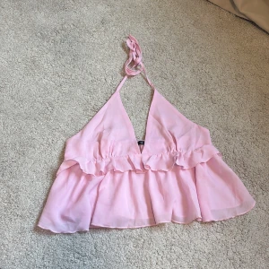 Ljusrosa volang topp med knyt från SHEIN - Säljer en ljusrosa, ärmlös topp med volangdetaljer och knyt i nacken. Toppen har v-ringning och är tillverkad i ett tunt, flowy material som ger en luftig känsla. Perfekt för varma dagar och somriga outfits.