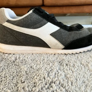 Svarta och grå sneakers från Diadora - Säljer ett par snygga Diadora sneakers i svart och grått med vit sula och vit sidodetalj. Skorna har snörning och är tillverkade i textil och syntetmaterial. Perfekta för en sportig och avslappnad stil.