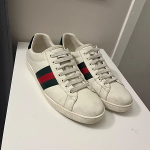 Vita sneakers från Gucci med röd och grön rand - Säljer ett par klassiska vita sneakers från Gucci med ikoniska ränder i grönt och rött på sidorna. Skorna har rund tå, platt sula och är tillverkade i skinn. Perfekta för dig som gillar stilrena och lyxiga sneakers.