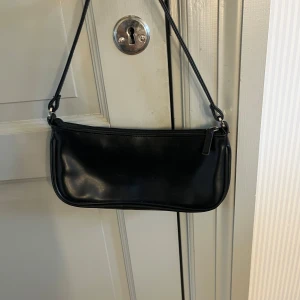 Svart axelväska  - Säljer en stilren svart axelväska i skinnimitation med dragkedja upptill. Väskan har en smal axelrem och minimalistisk design, perfekt för att bära det viktigaste. Enkel och snygg att matcha till olika outfits. 