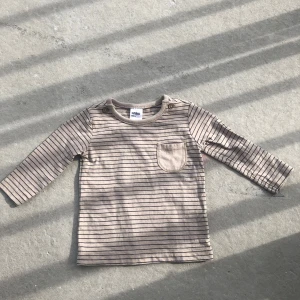 Beige och brunrandig långärmad tröja från Anko Baby - Säljer en beige långärmad tröja från Anko Baby med tunna bruna ränder och en liten bröstficka. Tröjan har knappar på axeln för enkel påklädning och är gjord i mjuk bomull. Perfekt för en stilren och bekväm look.