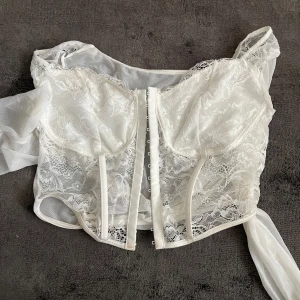 Vit spetskorsett med axelband - Säljer en vit korsettopp med spetsdetaljer och genomskinliga partier. Toppen har axelband och knäppning framtill. Perfekt för att skapa en romantisk och trendig look.