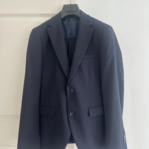 Mörkblå kostym från Riley - Säljer en stilren mörkblå kostym från Riley i storlek 48. Kostymen består av en kavaj med två knappar och matchande byxor. Materialet är en blandning av polyester, viskos och elastan för en bekväm passform. Använd 2 gånger, fläck fri. 