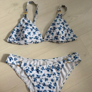 Vit bikini med blå blommor och gulddetaljer - Superfin bikini i vitt med blått blommönster. Triangelformad överdel med justerbara axelband och små guldfärgade ringar som detalj. Underdelen har samma blommiga print och matchande gulddetaljer i sidorna lågmdifjade. Perfekt för stranden eller poolen! Båda i storlek S men skulle mer säga XS! Ändast provade!