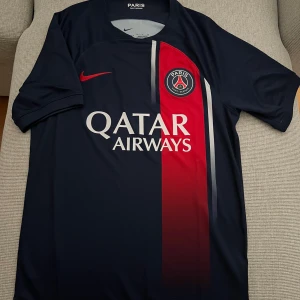 PSG Nike fotbollströja Dri-FIT S - Paris Saint-Germain matchtröja från Nike i Dri-FIT-material. Marinblå bas med röd vertikal rand och vita detaljer, PSG-emblem på bröstet och Qatar Airways-tryck. Klassisk rund hals och korta ärmar.