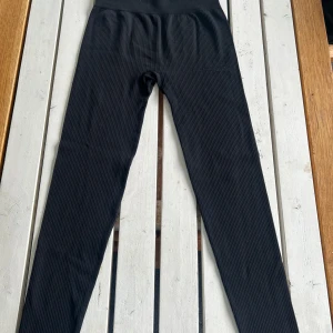 Svarta ribbade leggings New Apparel - Svarta ribbade leggings från New Apparel i storlek S. Hög midja och stretchigt material som sitter tajt och bekvämt. Diskret logga på låret och bred linning för extra stöd. (Använd enstaka gång)