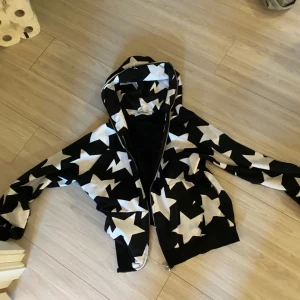 BAPE svart hoodie med stjärnor  - Svart hoodie från A Bathing Ape med stora vita stjärnor över hela jackan. Jackan har dragkedja framtill, huva och ribbade muddar. Perfekt för dig som gillar streetwear och vill sticka ut med ett unikt mönster. Står S men passar M 