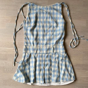 Blåvit rutig kortklänning NS76 - Supersöt kortklänning i blåvitt rutigt från NS76. Klänningen har knytband vid axlarna, markerad midja och plisserad kjol. Perfekt för sommarens alla tillfällen och ger en fräsch och ungdomlig look. Materialet känns mjukt och lätt mot huden. Ovansidan går även att vika ner för ytterliggare en look.